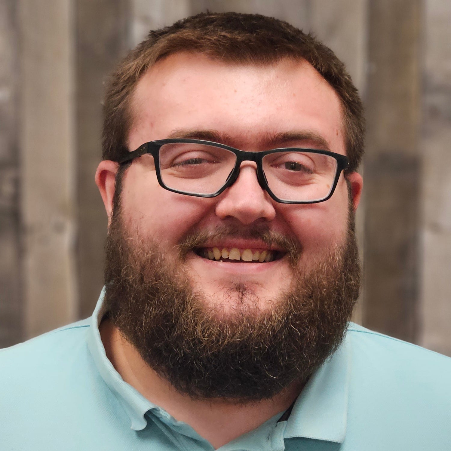 Man wearing glasses and a light blue shirt with a logo, standing against a wooden background